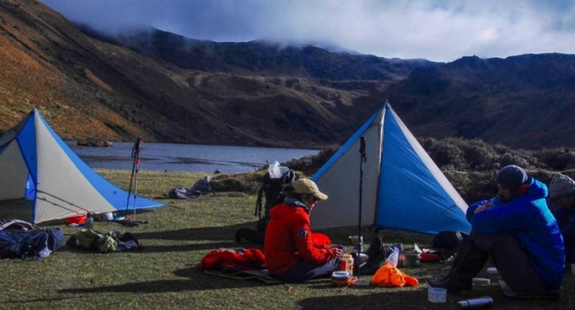 Two tents sit in an open area beside an alpine lake. There are mountains surrounding the lake.